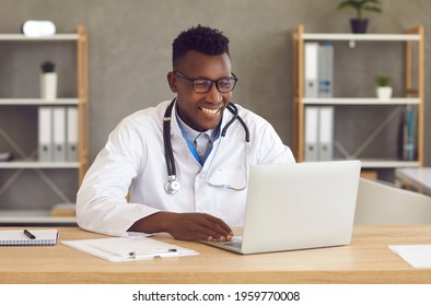 Happy medical specialist working on computer. Handsome black doctor sitting at office desk and looking at laptop screen during webcam video call with patient, or online educational training or webinar