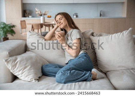 Happy merry middle-aged European woman sitting on comfortable sofa with eyes closed, dreaming, thinking of pleasant things, drinking hot tea, coffee, cocoa, smelling aroma, relaxing in cozy atmosphere