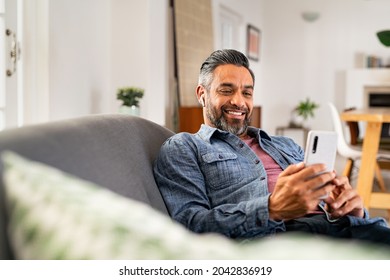 Happy mid adult man using smartphone device while sitting on sofa at home. Smiling mature indian man lying on couch reading messages on mobile phone while listening to music with wireless earbuds.