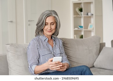 Happy Mid Age Senior Adult Woman Using Smartphone Sitting On Sofa At Home. Relaxed Mature Older Lady Holding Mobile Phone Texting, Buying Online, Checking Apps On Cellphone Tech Device On Couch.