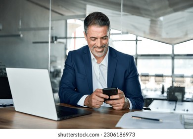 Happy mid aged business man ceo executive, financial expert investor wearing suit sitting in modern office holding smartphone using mobile phone managing digital tech work transactions at workplace. - Powered by Shutterstock