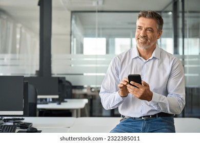 Happy mid aged business man using phone in office. Smiling mature confident businessman executive manager holding smartphone working on cell phone sitting at office desk looking away and thinking. - Powered by Shutterstock