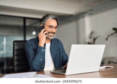 Happy mid aged business man manager talking consulting client on cell phone working on laptop in office. Busy smiling mature professional businessman making call on cell using computer sitting at desk - Powered by Shutterstock