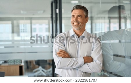 Happy mid aged older professional business man, mature latin executive manager, 50 years old investor standing in office arms crossed leaning at glass wall looking away thinking of success concept.
