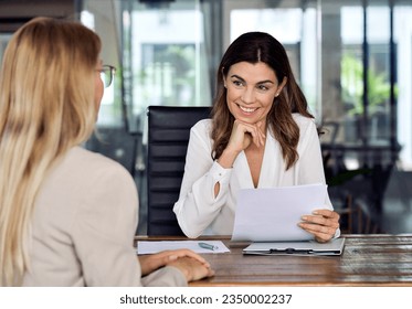 Happy mid aged professional business woman executive hr manager having job interview or business discussion with female applicant holding cv sitting at workplace in corporate office meeting.
