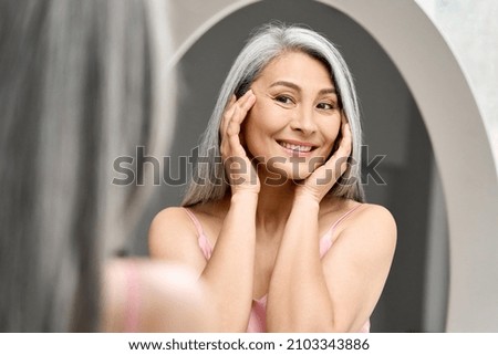 Happy middle 50 years aged asian woman with gray hair looking at mirror reflection examining touching face enjoying antiaging beauty treatments. Beauty hydrate skin care wrinkle prevention concept.