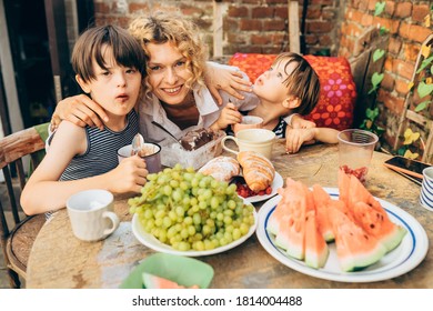 Happy Middle Age Blond Mother Carring Her Children. Mature Woman With Sons Eating Fruits And Dessert At Terrace Over Brick Wall On Backyard. Childern Need Care, Presence And Love.