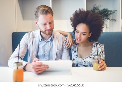 Happy Middle Age Interracial Couple Sitting In Cafe Bar, Smiling And Looking At White Tablet.