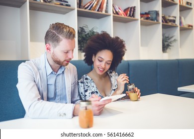 Happy Middle Age Interracial Couple Sitting In Cafe Bar, Smiling And Looking At White Tablet.