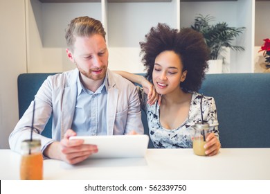 Happy Middle Age Interracial Couple Sitting In Cafe Bar, Smiling And Looking At White Tablet.