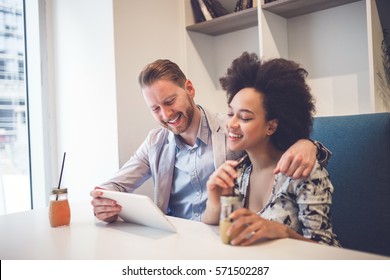 Happy Middle Age Interracial Couple Sitting In Cafe Bar, Smiling And Looking At White Tablet.