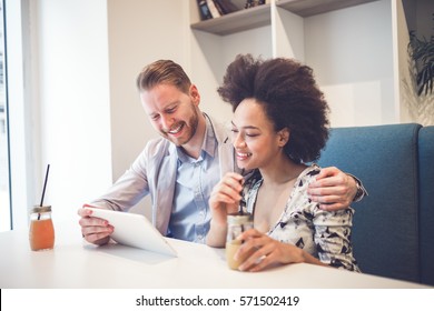 Happy Middle Age Interracial Couple Sitting In Cafe Bar, Smiling And Looking At White Tablet.