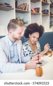 Happy Middle Age Interracial Couple Sitting In Cafe Bar, Smiling And Looking At White Tablet.