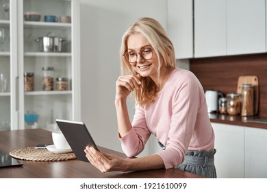 Happy Middle Aged 50 Years Old Woman Using Digital Tablet Sitting In Kitchen At Home. Mature Older Lady Wearing Glasses Holding Pad Computer Reading E Book, Shopping Online, Making Video Call.