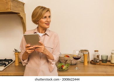 Happy Middle Aged 50 Years Old Woman Using Digital Tablet Sitting In Kitchen At Home. Mature Older Lady  Holding Pad Computer, Finding Recipe, Shopping Online, Making Video Call.