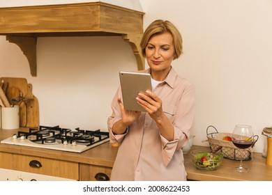 Happy Middle Aged 50 Years Old Woman Using Digital Tablet Sitting In Kitchen At Home. Mature Older Lady  Holding Pad Computer, Finding Recipe, Shopping Online, Making Video Call.