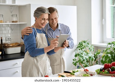 Happy Middle Aged 50s Couple Preparing Salad Using Digital Tablet In Kitchen. Smiling Mature Older Husband And Wife Holding Computer Cooking Together Searching Healthy Diet Food Online Recipes At Home