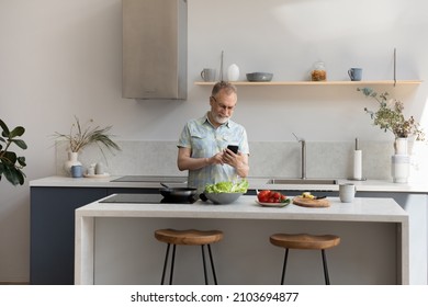 Happy Middle Aged 60s Man Distracted From Preparing Food Using Cellphone, Web Surfing Recipe For Tasty Meal, Communicating Distantly In Social Network While Cooking Vegetarian Dinner In Modern Kitchen