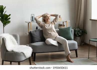 Happy Middle Aged 60s Pensioner Spending Leisure Time At Home, Sitting On Couch, Looking At Window, Relaxing In Modern Living Room. Elder Lady Enjoying Wealthy Retirement, Thinking Future Vacation