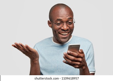 Happy Middle Aged African Amerfican Male Holds Modern Smart Phone, Recieves Good Message On Cellular, Reads News In Internet, Has Plesant Smile, Keeps Hand Raised, Poses Indoor. Technology Concept