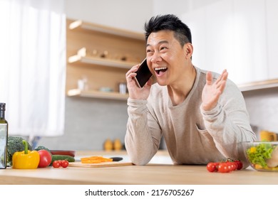 Happy Middle Aged Asian Man Having Phone Conversation While Cooking At Home, Looking At Copy Space And Gesturing, Inviting His Friends For Healthy Delicious Dinner, Kitchen Interior