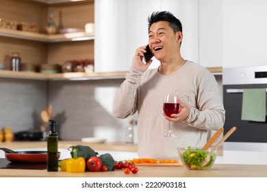 Happy Middle Aged Asian Man In Casual Outfit Standing Next To Kitchen Table At Home, Cooking Healthy Meal And Talking On Mobile Phone, Holding Glass Of Wine And Looking At Copy Space