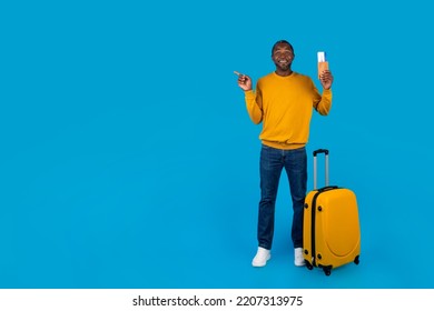 Happy Middle Aged Black Man In Casual Outfit Having Journey Alone, Holding Passport With Flight Tickets, Standing Next To Suitcase, Pointing At Copy Space, Blue Studio Background, Full Length