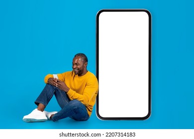 Happy middle aged black man in casual outfit sitting on floor next to huge cell phone with white empty screen over blue studio background, using smartphone and smiling, mockup, copy space - Powered by Shutterstock