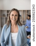 Happy middle aged business leader woman standing in office space with team working together, talking in background, looking at camera, smiling for portrait, enjoying leadership. Vertical head shot