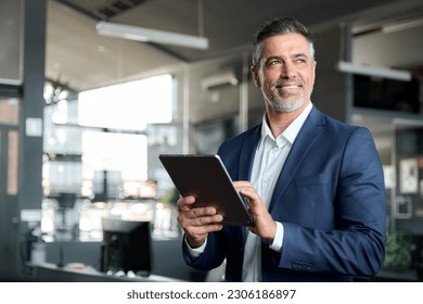 Happy middle aged business man ceo wearing suit standing in office using digital tablet. Smiling mature businessman professional executive manager looking away thinking working on tech device. - Shutterstock ID 2306186897