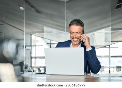 Happy middle aged business man talking on mobile phone working on laptop in office. Professional businessman executive wearing suit making call on cell using computer communicating with client. - Powered by Shutterstock