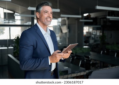 Happy middle aged business man ceo wearing suit standing in office using digital tablet. Smiling mature businessman professional executive manager looking away thinking working on fintech device. - Powered by Shutterstock