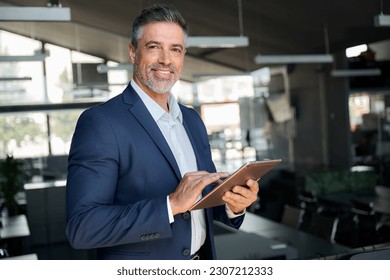 Happy middle aged business man ceo wearing suit standing in office using digital tablet. Confident mature businessman professional company executive looking at camera holding tech device, portrait.