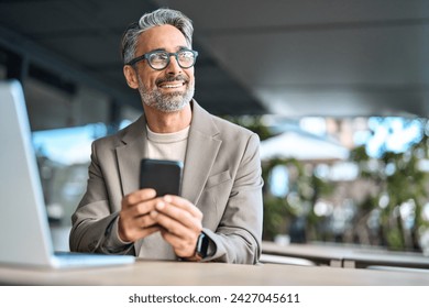 Happy middle aged business man executive using mobile phone sitting outside office. Stylish older busy businessman investor wearing glasses holding smartphone looking away at copy space with cellphone - Powered by Shutterstock
