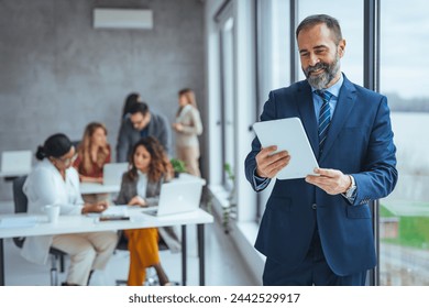 Happy middle aged business man ceo wearing suit standing in office using digital tablet. Smiling mature businessman professional executive manager looking away thinking working on tech device. - Powered by Shutterstock