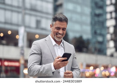 Happy middle aged business man wearing suit earbud holding phone having call walking on city street. Mature businessman with earphone using smartphone listening podcast in app on cellphone outdoors. - Powered by Shutterstock