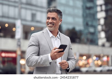 Happy middle aged business man wearing suit and earphone talking on mobile phone walking on busy city street. Mature businessman in earbuds making call or listening podcast outdoors. Authentic photo. - Powered by Shutterstock