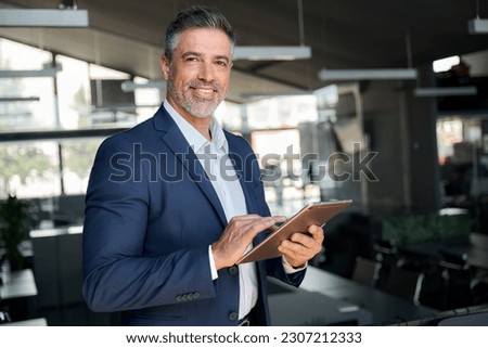 Happy middle aged business man ceo wearing suit standing in office using digital tablet. Confident mature businessman professional company executive looking at camera holding tech device, portrait.