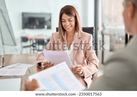 Happy middle aged business woman account manager discussing documents financial result project overview at office meeting, two professional executives holding tax papers consulting working together.