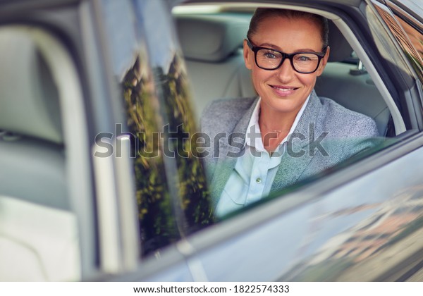 Happy middle aged business woman wearing eyeglasses\
looking out of a car window while sitting on back seat, she is\
going for a meeting by\
taxi