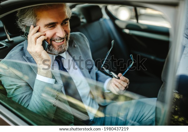 Happy middle aged businessman using\
cell phone in car, having phone call and holding\
glasses.