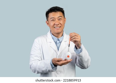 Happy Middle Aged Chinese Male Doctor In White Coat Showing Emergency Alarm Button Isolated On Blue Background, Studio. Professional Recommendation, Medicine, Health Care During Covid-19 Pandemic