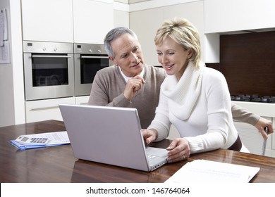 Happy Middle Aged Couple Calculating Home Finances On Laptop At Kitchen Table