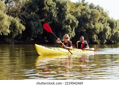 Happy Middle Aged Couple Kayaking On Yellow Kayak In Summer River Together. Having Fun In Leisure Activity. Romantic And Happy Woman And Man On The Kayak. Sport, Relations Concept.