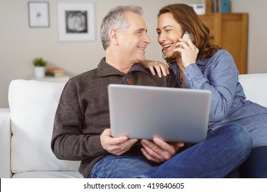 Happy Middle Aged Couple Seated On White Couch In Their Living Room With Laptop While The Wife Makes A Phone Call