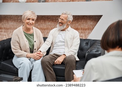 happy middle aged couple sitting on leather couch and holding hands near family counselor - Powered by Shutterstock