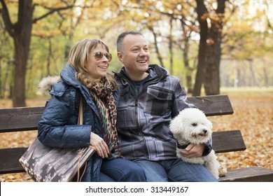 Happy Middle Aged Couple Sitting On Park Bench With Their Dog And Smiling.