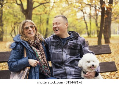 Happy Middle Aged Couple Sitting On Park Bench With Their Dog And Smiling.