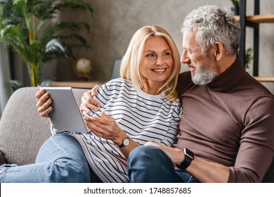Happy middle aged couple using digital tablet on sofa at home while looking on each other