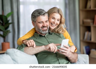 Happy Middle Aged Couple Using Mobile Phone At Home Together, Browsing Internet Or Shopping Online, Husband Pointing Finger At Smartphone Screen, Showing Something To His Smiling Wife, Closeup